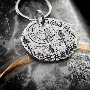 May include: A silver keychain with a circular charm featuring a crescent moon, two figures standing under the moon, a mountain range, and trees. The charm is engraved with the text "3,653 Moons" and "05.15.15 C+A".
