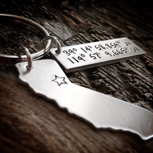 May include: Silver keychain with a small silver tag engraved with the coordinates "39.0 119.0 14.58.8591 34.57.9.6665 W". The keychain also features a silver outline of the state of California with a star in the center.