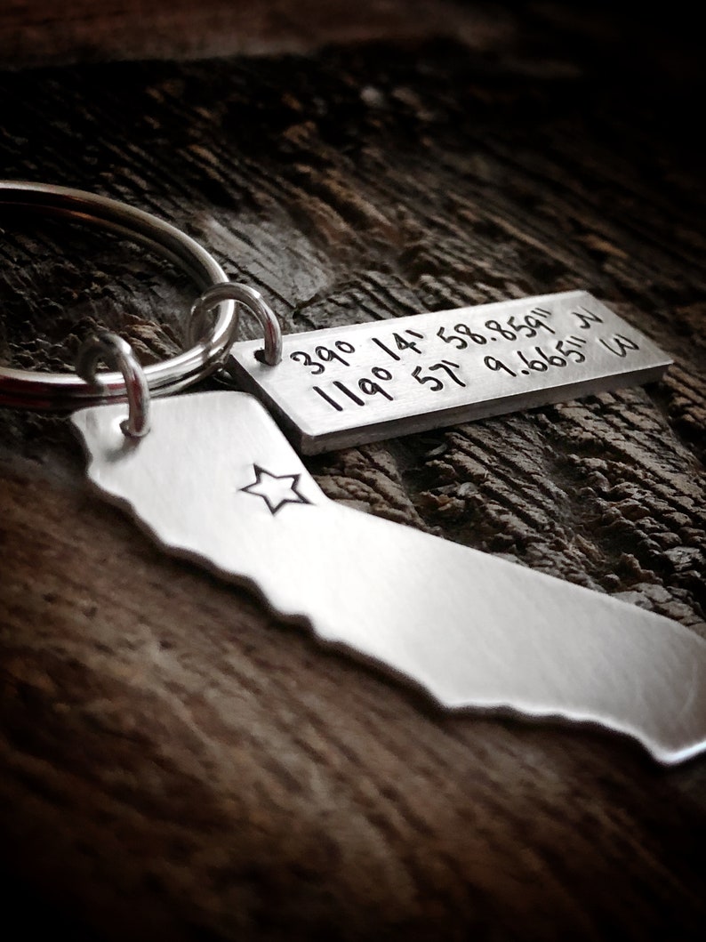 May include: Silver keychain with a small silver tag engraved with the coordinates "39.0 119.0 14.58.8591 34.57.9.6665 W". The keychain also features a silver outline of the state of California with a star in the center.