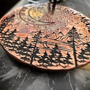 Op de afbeelding: Ronde koperen hanger met een gedetailleerd ontwerp van een halve maan, sterren, bergen en een bos met bomen. De hanger heeft een rustieke, vintage uitstraling.
