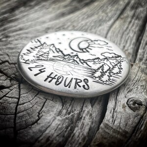 Peut inclure: Un disque rond en argent avec un motif grav&eacute; repr&eacute;sentant une cha&icirc;ne de montagnes, des arbres, une lune et un soleil. Le texte "24 HOURS" est grav&eacute; sur le disque.