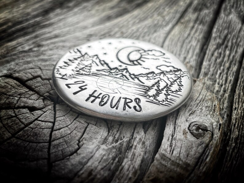 Peut inclure: Un disque rond en argent avec un motif grav&eacute; repr&eacute;sentant une cha&icirc;ne de montagnes, des arbres, une lune et un soleil. Le texte "24 HOURS" est grav&eacute; sur le disque.