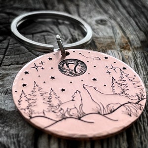 Puede incluir: Un llavero redondo de color cobre con un dise&ntilde;o de lobo aullando a la luna. El llavero tiene un anillo de llave plateado y un peque&ntilde;o c&iacute;rculo grabado con el n&uacute;mero "7" en su interior. El dise&ntilde;o tambi&eacute;n incluye un cielo estrellado, pinos y una luna creciente.
