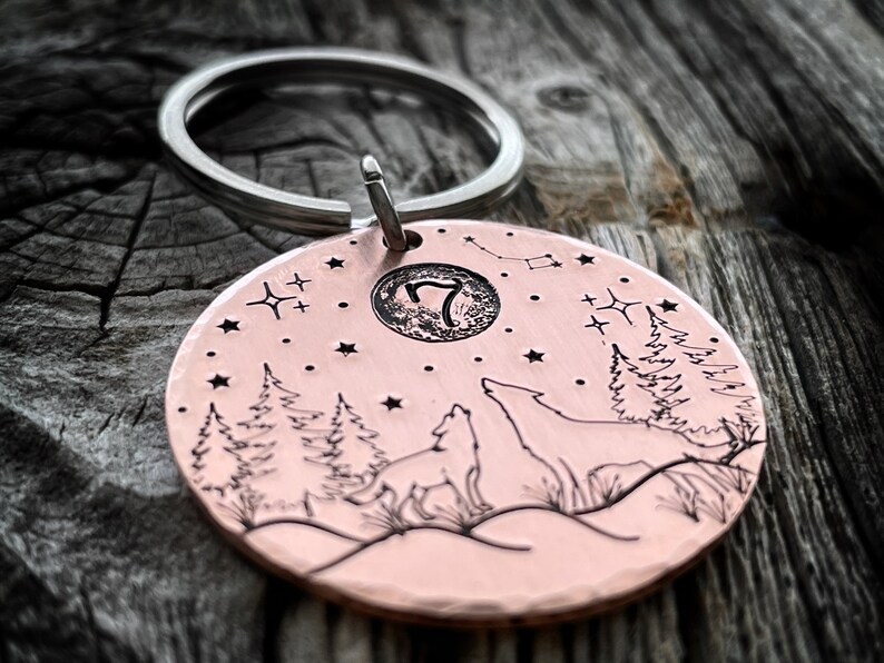 Puede incluir: Un llavero redondo de color cobre con un dise&ntilde;o de lobo aullando a la luna. El llavero tiene un anillo de llave plateado y un peque&ntilde;o c&iacute;rculo grabado con el n&uacute;mero "7" en su interior. El dise&ntilde;o tambi&eacute;n incluye un cielo estrellado, pinos y una luna creciente.