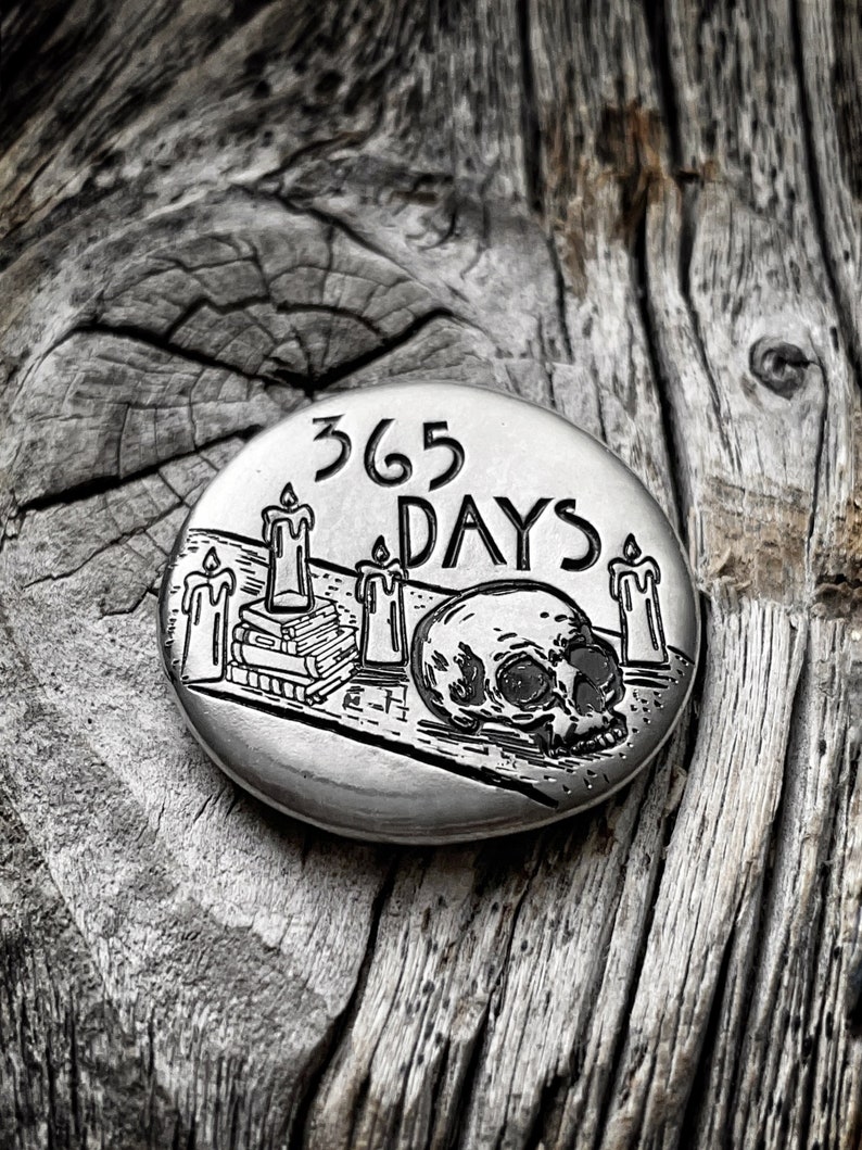 May include: A silver round metal token with a skull and crossbones design. The token has the text "365 DAYS" engraved on it.