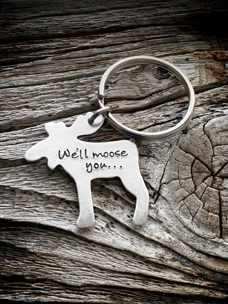 以下が含まれることがあります： 「Well moose you...」の文字が刻まれた、シルバーカラーのヘラジカ型キーホルダー。キーホルダーはシルバーのキーリングに付いています。背景は、風化した木材の質感のある表面です。
