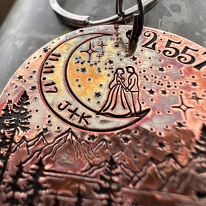 Op de afbeelding: Een koperkleurige metalen sleutelhanger met een halve maan ontwerp, een stel dat op de maan staat en een bergketen op de achtergrond. De sleutelhanger heeft ook een datum, "2.5.51", en de initialen "J+K".