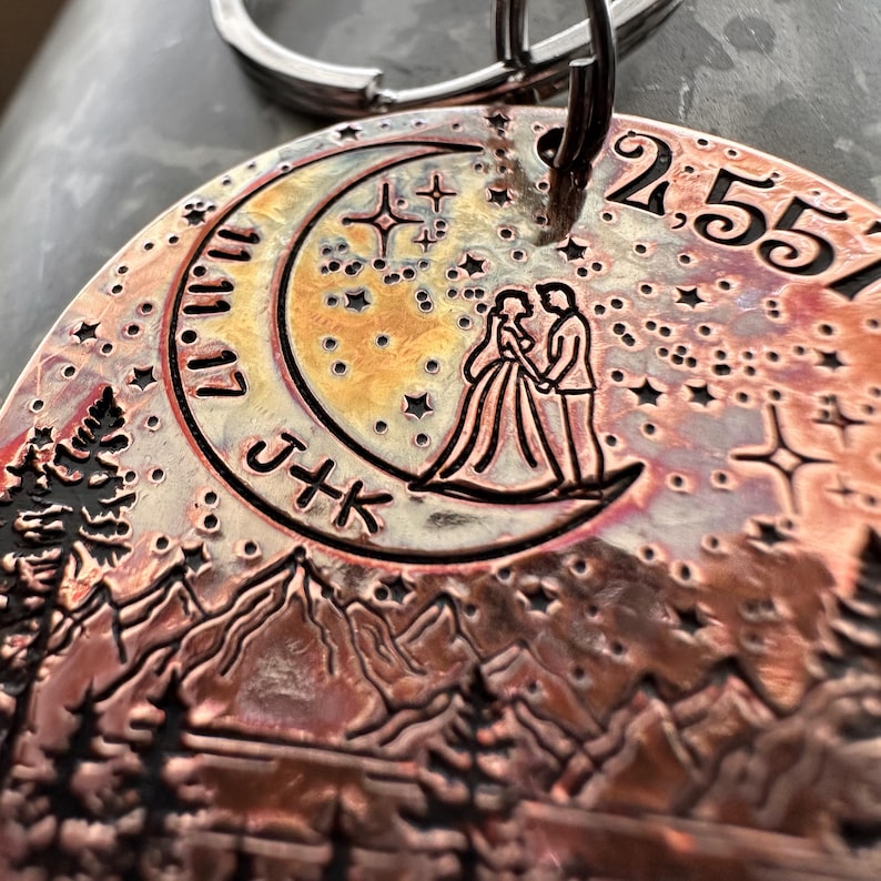 Op de afbeelding: Een koperkleurige metalen sleutelhanger met een halve maan ontwerp, een stel dat op de maan staat en een bergketen op de achtergrond. De sleutelhanger heeft ook een datum, "2.5.51", en de initialen "J+K".