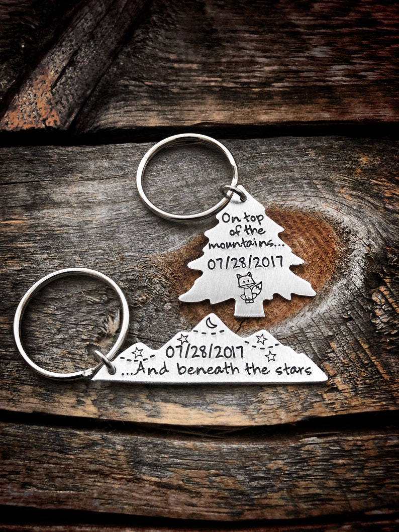 May include: Two silver keychains with a mountain and tree design. The mountain keychain says "...And beneath the stars" and "07/28/2017". The tree keychain says "On top of the mountains..." and "07/28/2017".
