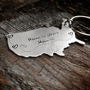 Puede incluir: Un llavero de metal plateado con forma de Estados Unidos con el texto "Home is where Mom is." y una l&iacute;nea punteada con corazones en la parte inferior.
