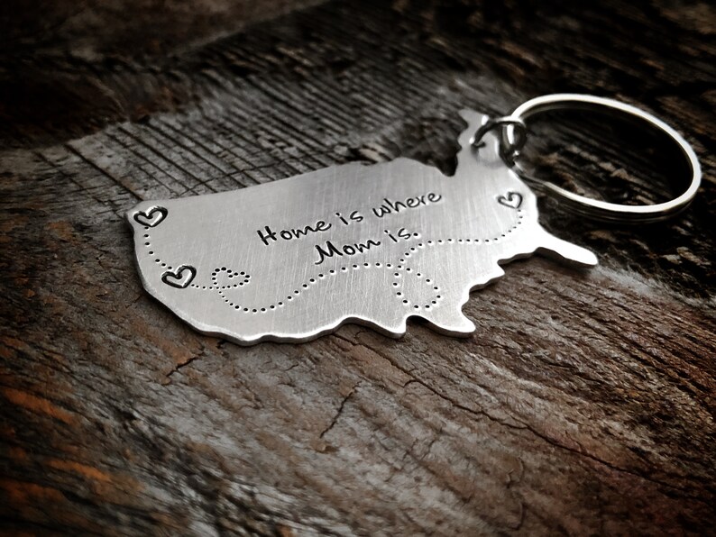 Puede incluir: Un llavero de metal plateado con forma de Estados Unidos con el texto "Home is where Mom is." y una l&iacute;nea punteada con corazones en la parte inferior.