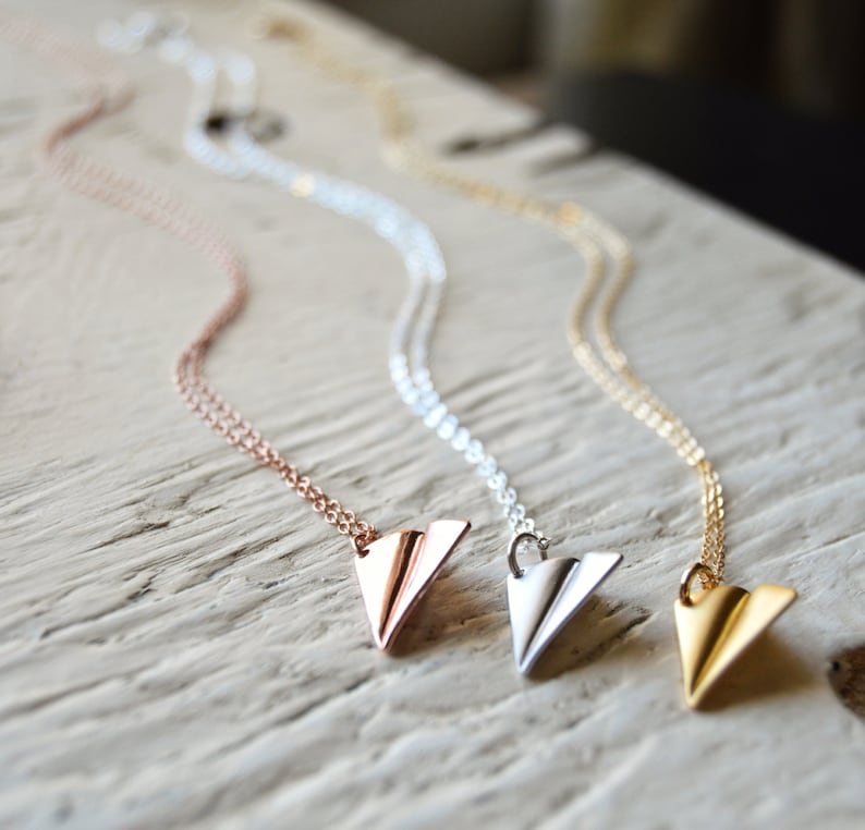 May include: Three necklaces with paper airplane pendants in rose gold, silver, and gold. The pendants are folded with detailed lines, and the chains are delicate. The necklaces are displayed on a light-colored, textured surface.