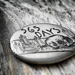 May include: A silver-toned metal coin with a skull and candles engraved on it. The coin reads "365 DAYS".