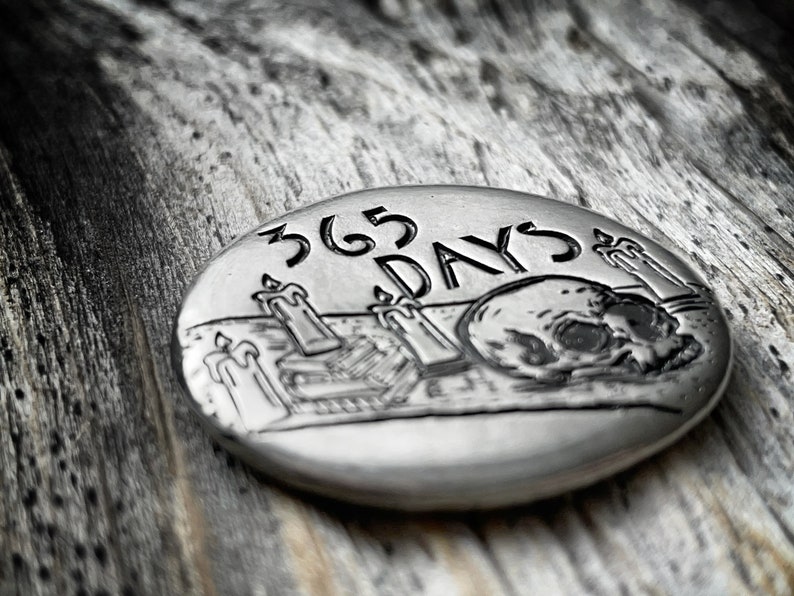 May include: A silver-toned metal coin with a skull and candles engraved on it. The coin reads "365 DAYS".