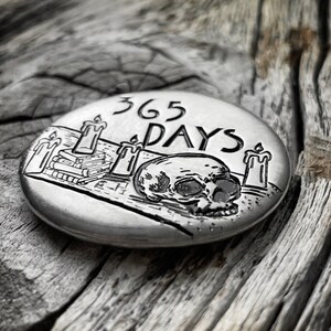 May include: A silver-toned round metal disc with an engraved design of a skull, three candles, and a stack of books. The text "365 DAYS" is engraved above the skull.