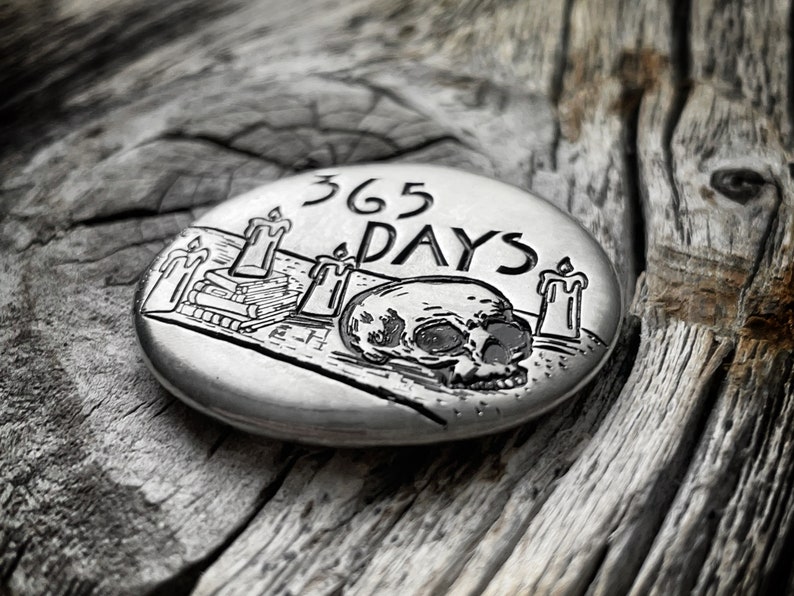 May include: A silver-toned round metal disc with an engraved design of a skull, three candles, and a stack of books. The text "365 DAYS" is engraved above the skull.