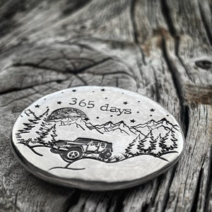 May include: A silver round disc with an engraved design of a jeep driving through a mountain landscape with a full moon and stars. The text "365 days" is engraved above the jeep.