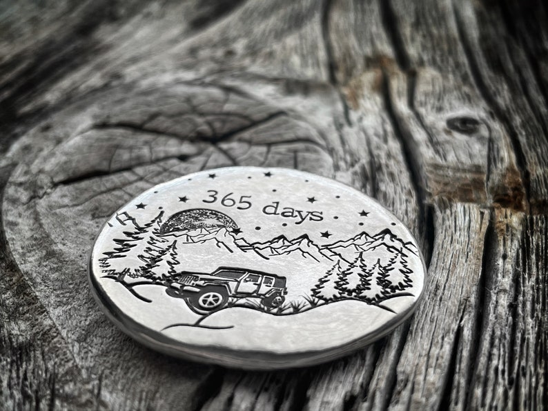May include: A silver round disc with an engraved design of a jeep driving through a mountain landscape with a full moon and stars. The text "365 days" is engraved above the jeep.