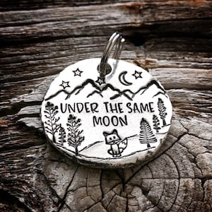 May include: Silver charm with a mountain scene, a crescent moon, stars, and a fox. The charm says "Under the Same Moon".
