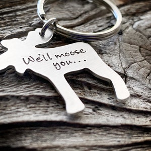 以下が含まれることがあります： 「We'll moose you...」の文字が刻まれた、シルバーカラーのヘラジカ型キーホルダー。キーホルダーはシルバーのリングに付いています。背景は風化した木製の表面です。