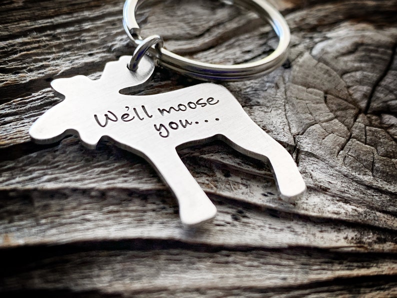 以下が含まれることがあります： 「We'll moose you...」の文字が刻まれた、シルバーカラーのヘラジカ型キーホルダー。キーホルダーはシルバーのリングに付いています。背景は風化した木製の表面です。
