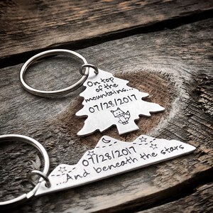 May include: A silver keychain with two charms. The first charm is a tree with the text "On top of the mountains... 07/28/2017". The second charm is a mountain range with the text "07/28/2017... And beneath the stars".