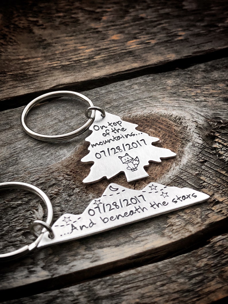 May include: A silver keychain with two charms. The first charm is a tree with the text "On top of the mountains... 07/28/2017". The second charm is a mountain range with the text "07/28/2017... And beneath the stars".