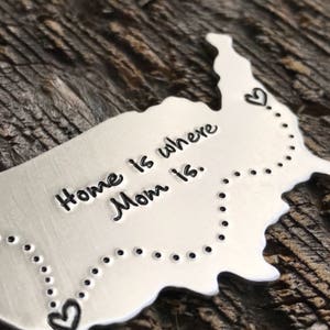 Puede incluir: Un contorno de metal plateado de los Estados Unidos con una l&iacute;nea punteada y corazones que conectan los bordes. El texto "Home is where Mom is." est&aacute; grabado en el contorno.