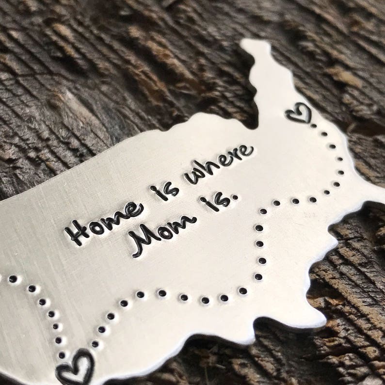 Puede incluir: Un contorno de metal plateado de los Estados Unidos con una l&iacute;nea punteada y corazones que conectan los bordes. El texto "Home is where Mom is." est&aacute; grabado en el contorno.