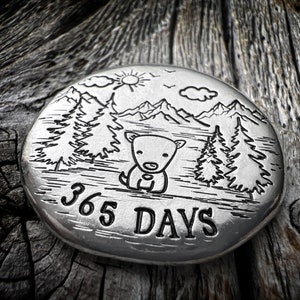 May include: A round, silver-colored metal pendant with an engraved design. The design features a cartoon dog, mountains, trees, sun, and the text "365 DAYS". The pendant is set against a rustic wooden background.