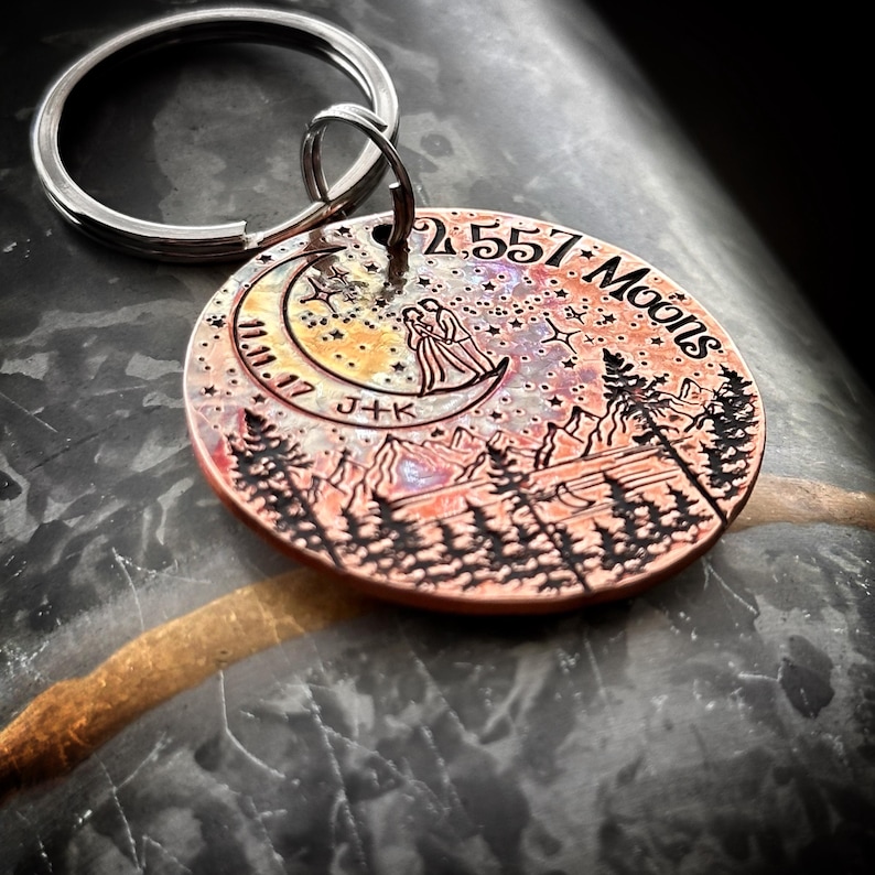 Op de afbeelding: Een koperkleurige sleutelhanger met een halve maan ontwerp, twee figuren die op de maan staan, en de tekst "2.557 Manen" en "11.10.17 J+K". De sleutelhanger toont een sterrenhemel en een bergketen op de achtergrond.