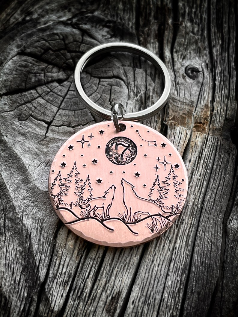 Puede incluir: Un llavero de color cobre con un dije redondo que presenta una luna, estrellas y dos lobos aullando a la luna. El dije est&aacute; grabado con el n&uacute;mero "7".