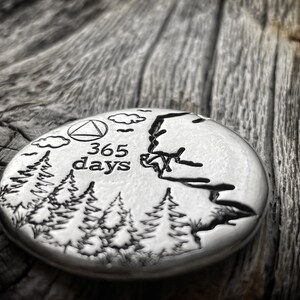 May include: A silver-colored metal token with a design of trees, clouds, and a triangle with the text "365 days" inside. The token is on a wooden surface.