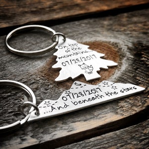 May include: Two silver keychains, one shaped like a pine tree and the other like a mountain range. The tree keychain has the text "On top of the mountains... 07/28/2017" and a small fox illustration. The mountain keychain has the text "...And beneath the stars 07/28/2017".