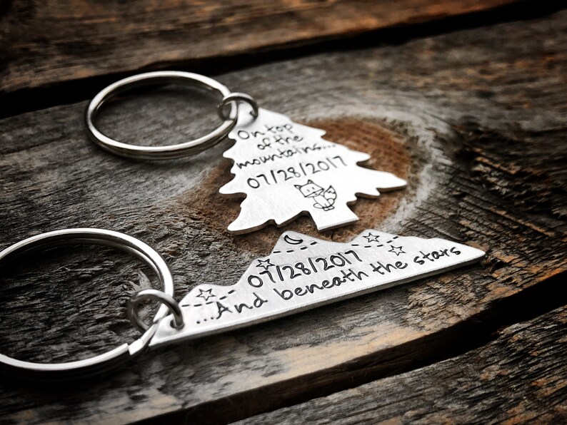 May include: Two silver keychains, one shaped like a pine tree and the other like a mountain range. The tree keychain has the text "On top of the mountains... 07/28/2017" and a small fox illustration. The mountain keychain has the text "...And beneath the stars 07/28/2017".