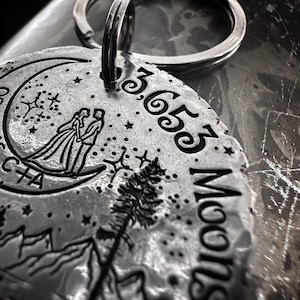 May include: A silver keychain with a crescent moon design, a couple standing on the moon, a pine tree, and mountains. The keychain also has the text "3,653 Moons" engraved on it.