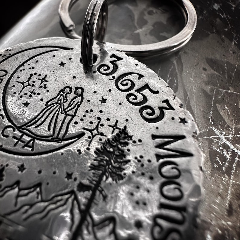 May include: A silver keychain with a crescent moon design, a couple standing on the moon, a pine tree, and mountains. The keychain also has the text "3,653 Moons" engraved on it.