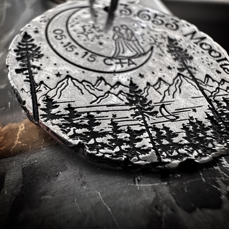 May include: A silver metal disc with a design of a mountain range, trees, a crescent moon, and a couple standing under a crescent moon. The text "05.15.15" and "3,650 Moons" are engraved on the disc.