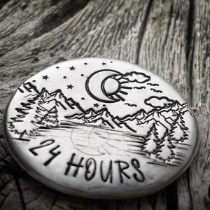 Peut inclure: Un disque rond en argent avec un motif de croissant de lune, de soleil, de montagnes et d'arbres. Le texte "24 HOURS" est grav&eacute; au bas du disque.