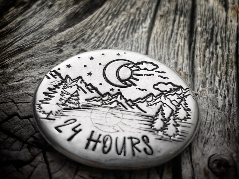 Peut inclure: Un disque rond en argent avec un motif de croissant de lune, de soleil, de montagnes et d'arbres. Le texte "24 HOURS" est grav&eacute; au bas du disque.