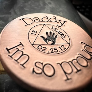K&ouml;nnte beinhalten: Eine runde kupferfarbene M&uuml;nze mit den Worten "Daddy" und "I'm so proud" eingraviert am Rand. In der Mitte befindet sich ein Dreieck mit den Worten "13 Years" und "02.25.12" darin.