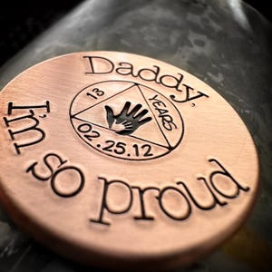 K&ouml;nnte beinhalten: Eine runde Scheibe aus Kupfer mit dem Text "Daddy, I'm so proud" eingraviert am Rand. In der Mitte befindet sich ein Dreieck mit dem Text "13 Years" und "02.25.12" im Inneren.