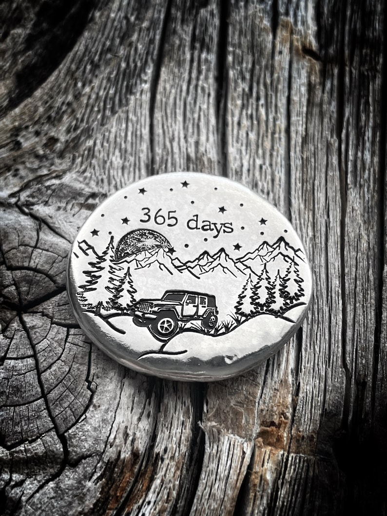 May include: A silver round metal disc with an engraved design of a jeep driving through a mountain landscape with trees and a moon. The text "365 days" is engraved on the disc.