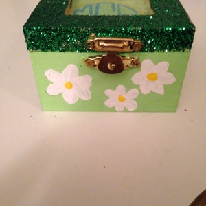 May include: A small green wooden box with a gold latch. The box is decorated with green glitter on the top and white flowers with yellow centres on the front. The box has a clear glass window on the top.
