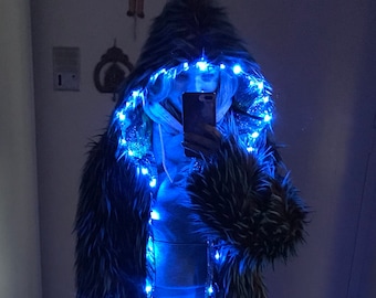 Puede incluir: Una larga capa de piel sintética negra y gris con capucha. La capa está decorada con luces LED azules.