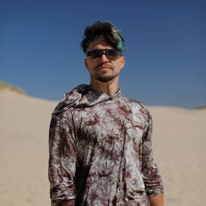 May include: A person wearing a brown and white tie-dye long-sleeve shirt with a hood. The shirt has a relaxed fit and is made of a soft, comfortable fabric. The person is also wearing blue jeans and a blue wristband.
