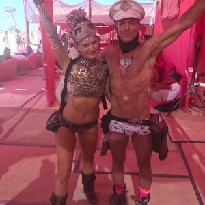 Puede incluir: Una pareja vestida con atuendos coloridos de festival. La mujer lleva un top de estampado de leopardo, pantalones cortos negros y plateados y botas negras. El hombre lleva un sombrero blanco, ropa interior blanca con estrellas y calcetines rosa y blancos. Ambos sonr&iacute;en y miran a la c&aacute;mara.