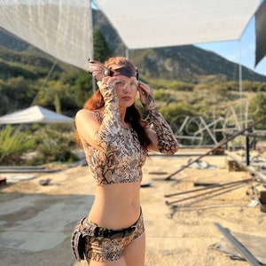 Puede incluir: Una mujer con un top corto y pantalones cortos a juego de estampado de serpiente marr&oacute;n y beige, con un cintur&oacute;n de cuero marr&oacute;n y una ri&ntilde;onera de cuero y metal marr&oacute;n. Lleva una diadema de cuero marr&oacute;n con un acento met&aacute;lico y gafas transparentes. La mujer est&aacute; de pie en un entorno des&eacute;rtico con una estructura blanca en el fondo.