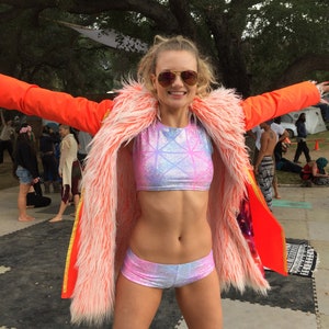 Puede incluir: Una mujer con una chaqueta de piel sint&eacute;tica rosa y blanca sobre un top corto rosa y azul y pantalones cortos a juego. Lleva gafas de sol y tiene el pelo recogido hacia atr&aacute;s.