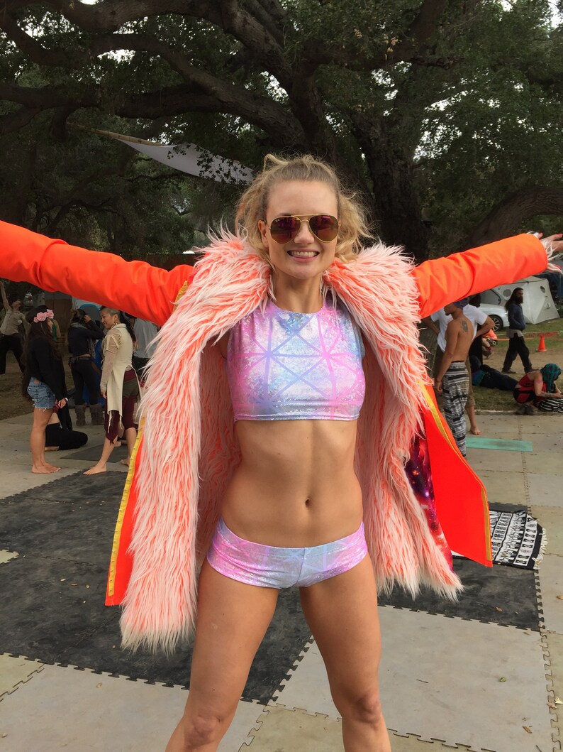 Puede incluir: Una mujer con una chaqueta de piel sint&eacute;tica rosa y blanca sobre un top corto rosa y azul y pantalones cortos a juego. Lleva gafas de sol y tiene el pelo recogido hacia atr&aacute;s.